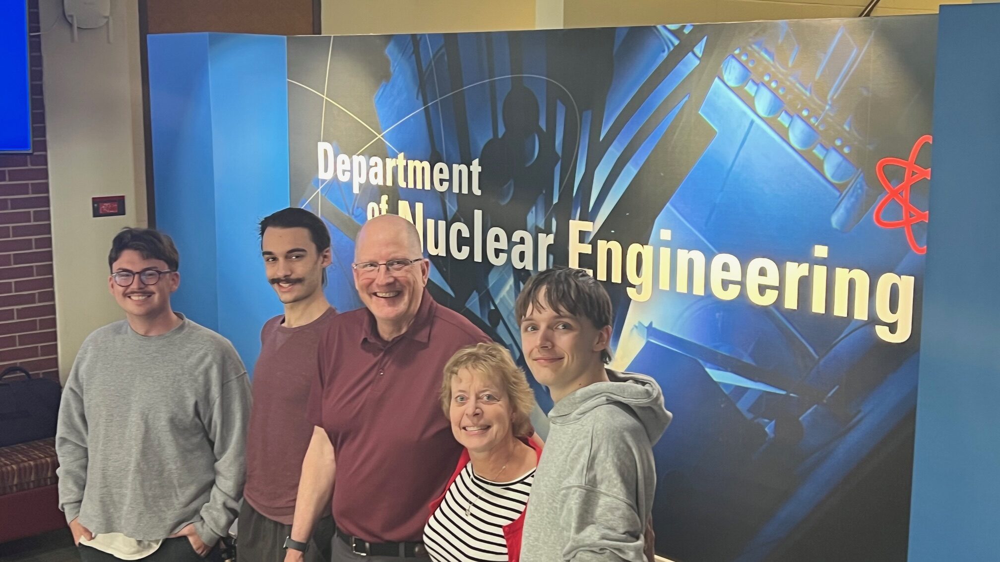 A group of people smiling in front of a sign that reads "Department of Nuclear Engineering.)