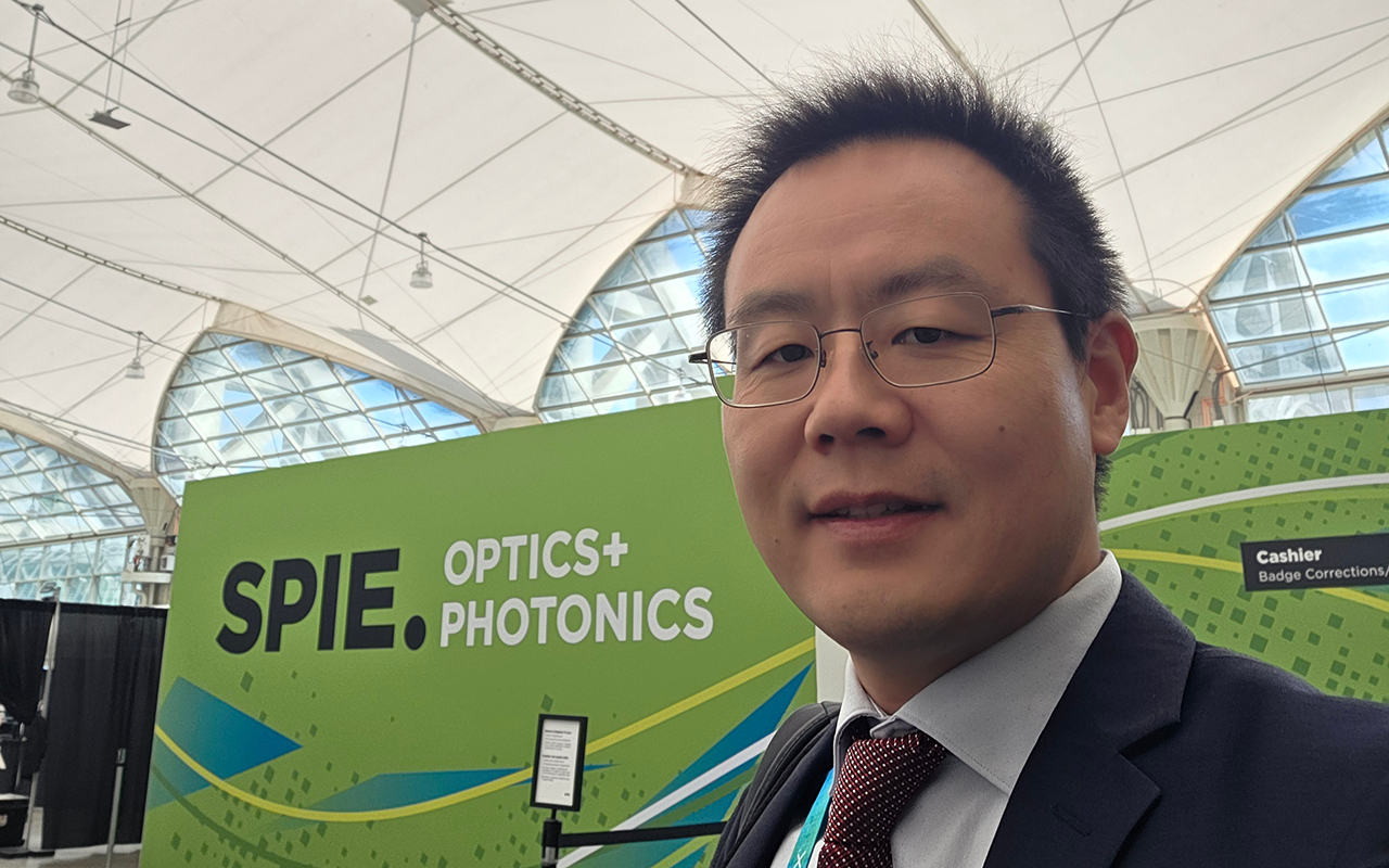A selfie of a man smiling with a green banner that reads "SPIE optics and photonics" behind him.